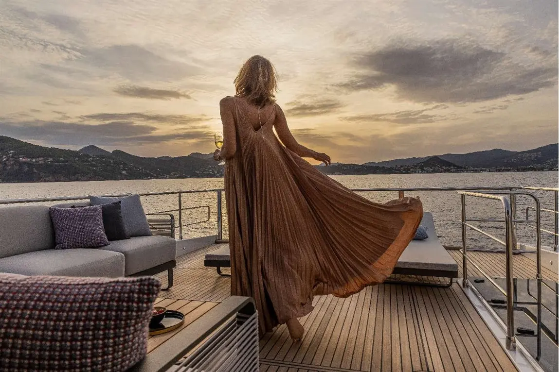 Woman in a flowing brown dress enjoys a sunset over the water, holding a drink on a yacht with lounge seating.